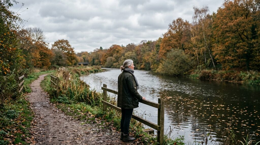 曇り空の川辺の道で、水面を見つめながら立ち止まる人物の後ろ姿と落ち葉が流れる風景